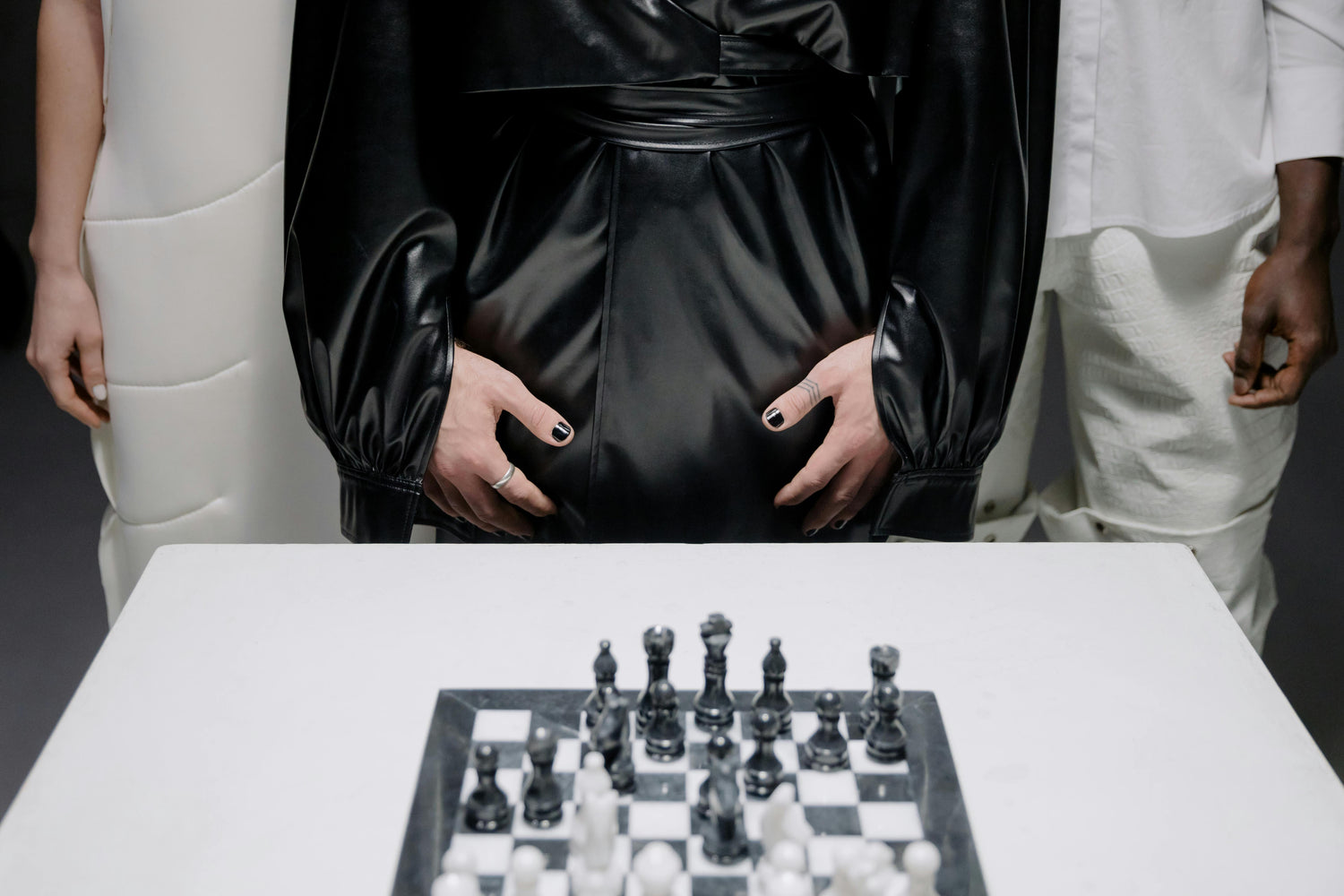 Person in a black dress standing behind a chessboard with another person in white partially visible.